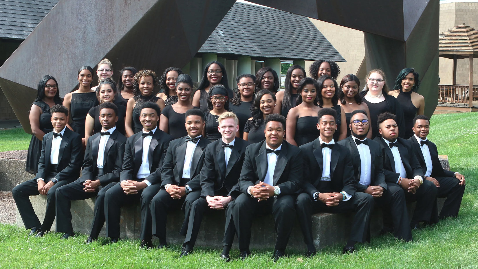 A large group of young adults dressed in formal black attire, with women in dresses and men in tuxedos, pose smiling together outdoors in front of a large abstract sculpture.